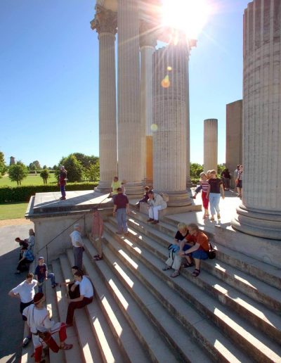 Das Foto zeigt einen Ausschnitt der Teilrekonstruktion des römischen Hafentempels im LVR-Archäologischen Park. Menschen halten sich bei Sonnenlicht auf der Treppe und zwischen den Säulen auf.